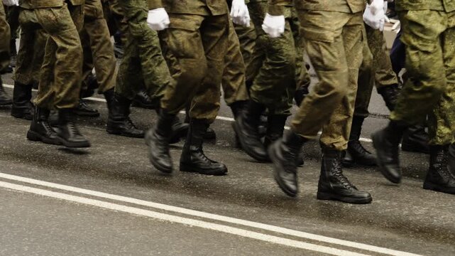 Close-up of synchronized soldiers boots and camouflage trousers marching in tight formation on a rain-soaked city street, with barriers and spectators along the wet roadway
