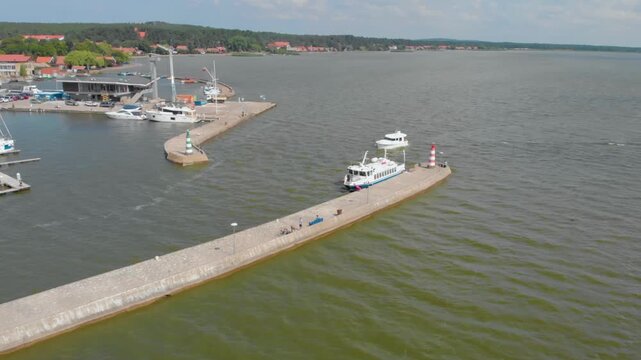 Drone footage captures the serene beauty of the Nida marina and the vast Curonian Lagoon in Lithuania. The shot showcases boats docked at the concrete pier, a small lighthouse, and the picturesque vil