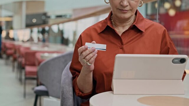 Cropped slowmo shot of adult woman sitting at table in modern mall and using credit card while making online payment on digital tablet