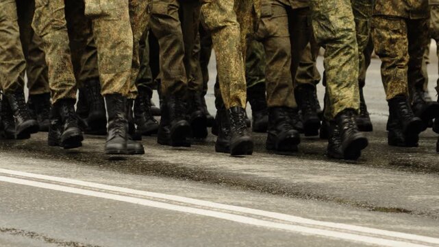 Close-up of uniformed soldiers in camouflage marching in formation during a disciplined parade, black combat boots striking the asphalt in synchronized steps along marked roadway