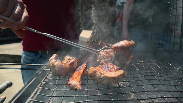 Close Up of Unrecognizable Cook Turning Grilled Pork Meat with Metal Tongs on Charcoal Barbecue Grill as Camera Slides Left and Right Through Smoke in Sunny Backyard Garden Summer Outdoor Cookout