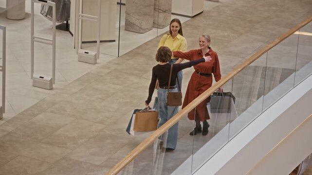 High angle full shot of three stylish Caucasian women carrying shopping bags meeting each other in modern mall and feeling happy to continue shopping together