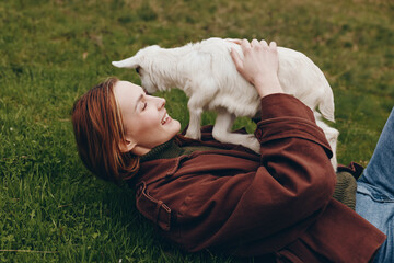 Fototapeta premium A young woman with red hair lies on grass smiling as she holds a white baby goat close to her face in a casual outdoor lifestyle moment.