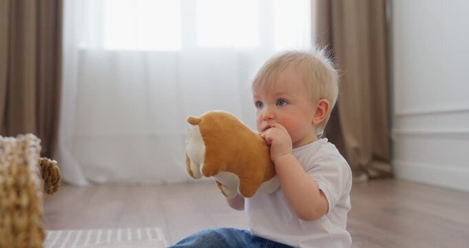 Blond toddler sitting on a wooden floor in a bright home interior, cuddling and chewing a small plush toy beside a woven basket and curtains