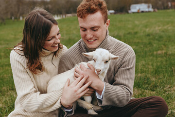 Fototapeta premium Lifestyle scene of a couple cuddling a goat kid in a field, wearing knit sweaters, they share affectionate smiles under sunlit grass, warm, wholesome moment in a rustic meadow