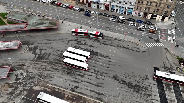 High-angle aerial drone footage of a bustling urban bus terminal with multiple buses maneuvering and vehicle traffic on an adjacent city street, symbolizing public transit and infrastructure.