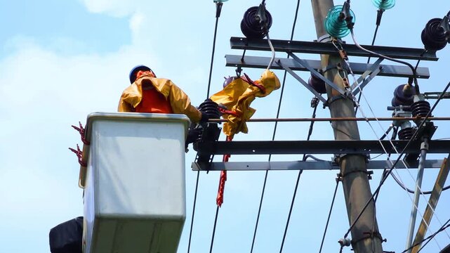 Electrician works on a tall pole. He wears bright yellow safety gear. He stands in a bucket lift, high above ground. The sky is blue with faint white clouds. Power lines and transformers hang nearby