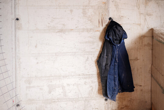 An old, weathered blue work jacket hangs on a hook against a large, rough concrete wall, providing ample copy space on the left for text or design elements.