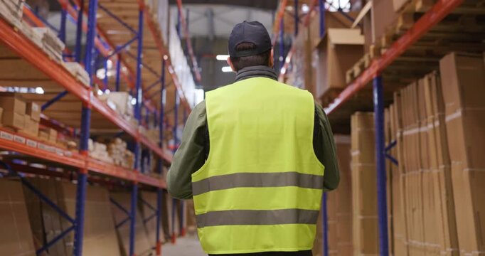 Warehouse worker inventory shelves logistics stock. Rear view of staff in vest checking goods on racking, managing stock levels for shipping needs. Clear concept of warehouse efficiency.