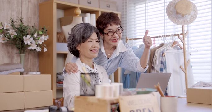 Adult asian daughter cheering beside senior mother at desk with laptop, both smiling in slowmotion, teamwork supporting family boutique fashion business success with online shop and handmade textile