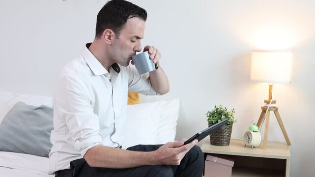 Businessman drinking coffee while using tablet in bedroom at home. Morning routine, remote work, digital lifestyle, online communication, and modern home office concept.