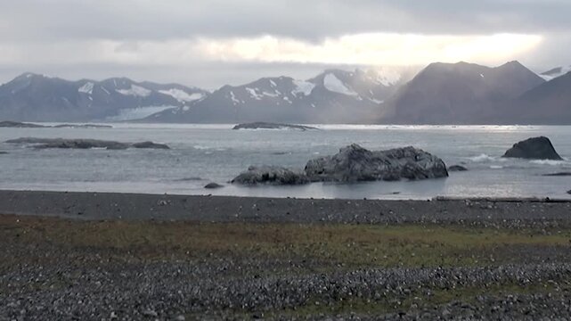 A vast panorama of Arctic Svalbard, Norway. Rugged, snow-dusted mountains loom over a restless ocean, its waves crashing against the pebble-strewn coast. A shy sunbeam graces the dramatic landscape.