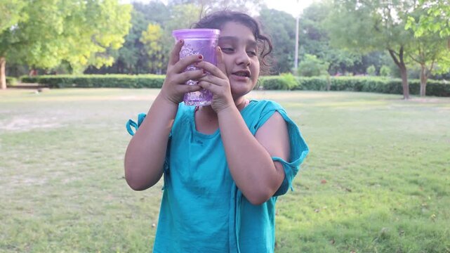 Young Indian girl holding a cold water bottle against her face while enjoying relief from hot summer weather in a green outdoor park. Refreshing lifestyle moment captured in India.
