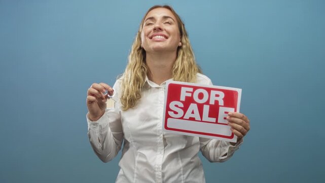 Woman holds keys and a for sale sign in studio, smiling while presenting keyring to camera; buying optimism.