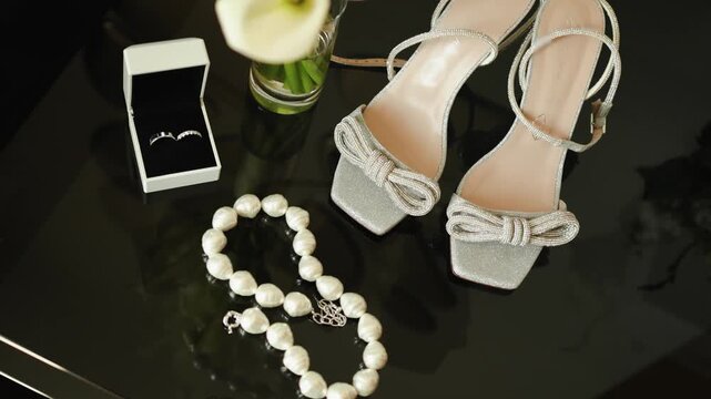 Wedding bouquet of white calla lilies flowers on table near elegant bride shoes with sparkling bow. Beautiful morning details concept with wedding rings in box, pearl necklace and jewelry layout in 4k