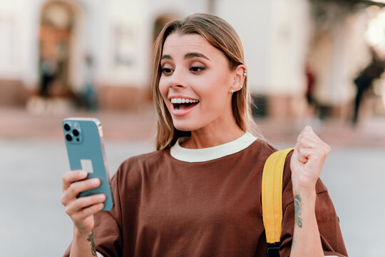 Happy young woman excitedly reading a message on her smartphone outdoors and celebrating good news while holding a yellow backpack strap in an urban street scene
