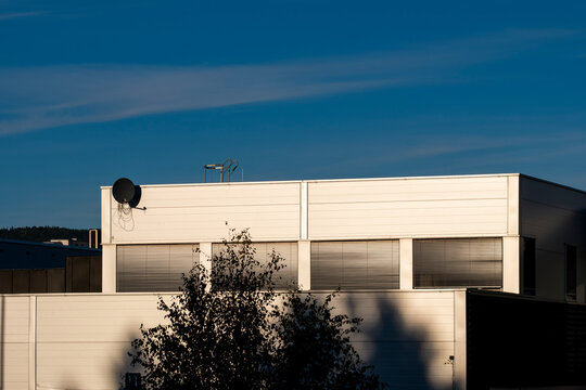 white rooftop wall catches warm shadow beneath blue sky as office facade, antenna, urban architecture and quiet building edge define the scene