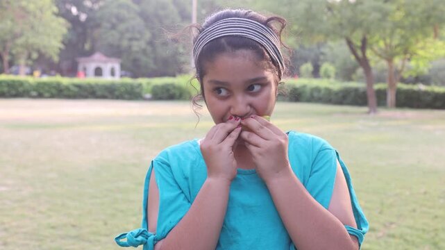  Young Indian girl eating fresh musk melon fruit while making a playful expression in a green park during hot summer weather. Cheerful outdoor lifestyle moment captured in natural surround