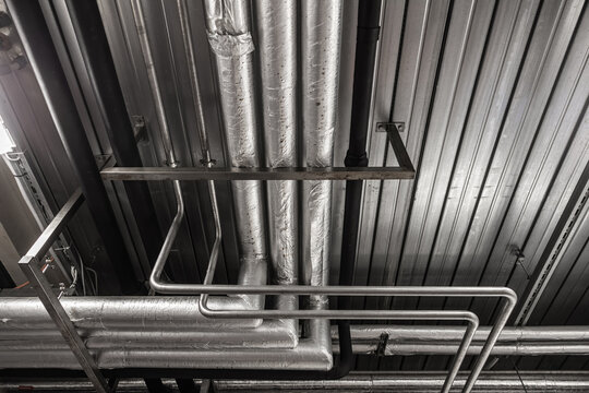 Ceiling view of insulated pipes, metal lines, support frames, and ducts inside a brewery or beverage production facility