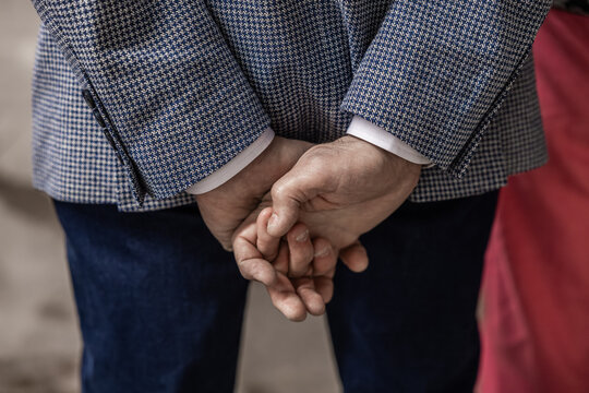 Close-up of a person clasping hands behind back while wearing a blue checkered blazer, with another figure in magenta clothing nearby at a social event