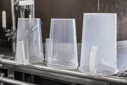 Translucent plastic measuring jugs upside down on stainless steel equipment inside a brewery or beverage production facility, with copy space