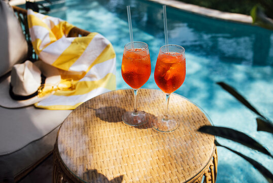 Two refreshing aperol spritz cocktails with orange slices and straws on a woven table beside a swimming pool, colorful towel draped over a lounge chair in the background