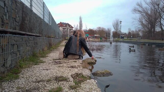 Child in a black leather jacket sits on a stone edge feeding mallard ducks in a Budapest park pond. A handheld wide shot showing the winter landscape, water fountain, and urban park environment.