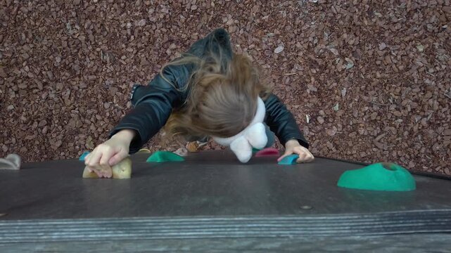 High angle static shot of a young girl wearing bunny earmuffs climbing a black outdoor rock wall. The background shows wood chips on the ground at the Varosliget park playground in Budapest Hungary.