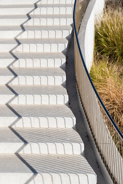 white stairs descend beside metal railing and dry grasses, where urban architecture, shadows and exterior steps shape a quiet public access path
