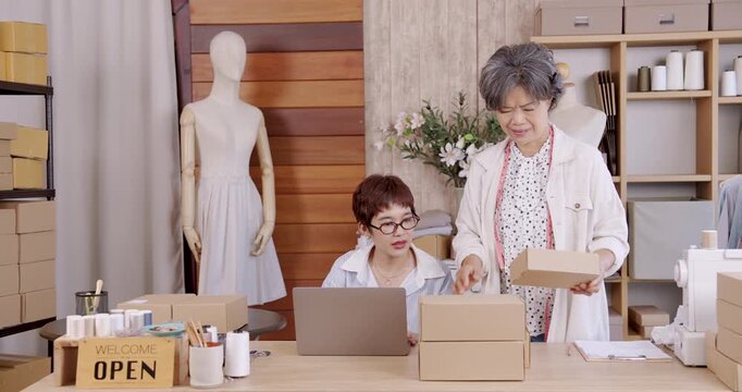 Senior asian mother holding cardboard box discussing with adult daughter working laptop inside boutique fashion studio family business teamwork ecommerce online shop packaging entrepreneurship