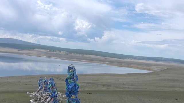 Experience the spiritual beauty of Mongolia as sacred Ovoo adorned with vibrant blue cloths stand sentinel by the shimmering waters of Lake Khuvsgul, a serene sight under a vast sky.