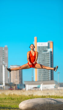 Athletic young woman in red bodysuit performing a high mid-air straddle split jump outdoors against modern city buildings.