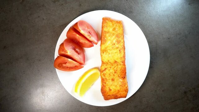 A high-angle close-up of a chef's hands plating a gourmet meal consisting of a seared fish fillet, fresh tomato slices, and a lemon wedge on a clean white plate.