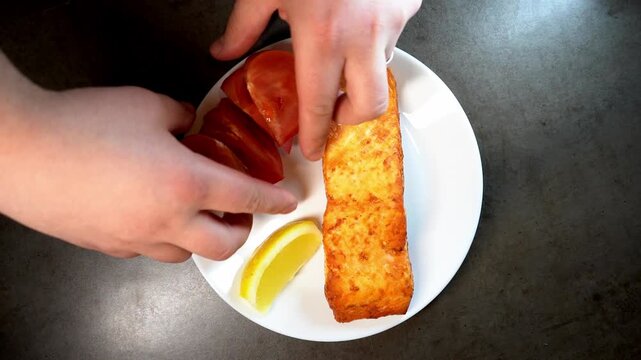 A high-angle close-up of a chef's hands plating a gourmet meal consisting of a seared fish fillet, fresh tomato slices, and a lemon wedge on a clean white plate.