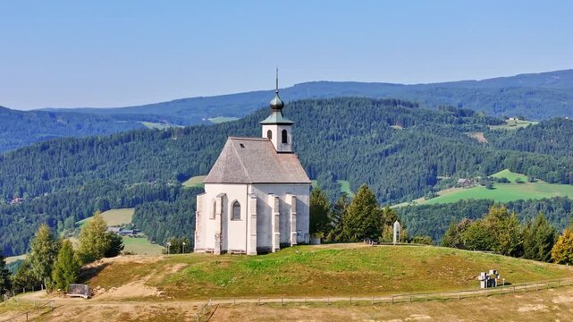 Aerial view of Wolfgangikirche, a white church on a grassy hilltop surrounded by dense green forests and rolling mountains under a clear blue sky, Schwanberg, Styria, Austria.