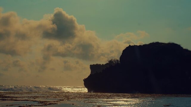 Golden sunlight illuminates crashing waves against a dark jagged cliffside on a serene beach at dawn with soft clouds overhead and a tranquil coastal atmosphere.
