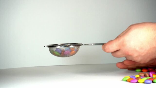 Close-up of a hand using a metal mesh strainer to scoop and manipulate colorful candy-coated chocolate drops on a clean white background, perfect for culinary and food-related concepts.