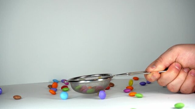 Close-up of a hand using a metal mesh strainer to scoop and manipulate colorful candy-coated chocolate drops on a clean white background, perfect for culinary and food-related concepts.