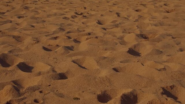 Textured ground-level view of a sandy surface featuring multiple indentations from footsteps, highlighting the natural grit, warm earth tones, and barren landscape aesthetic.