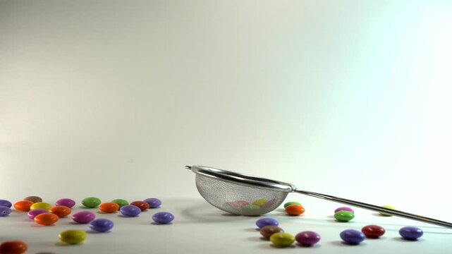 Close-up of a hand using a metal mesh strainer to scoop and manipulate colorful candy-coated chocolate drops on a clean white background, perfect for culinary and food-related concepts.