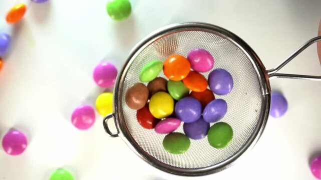 Close-up of a hand using a metal mesh strainer to scoop and manipulate colorful candy-coated chocolate drops on a clean white background, perfect for culinary and food-related concepts.