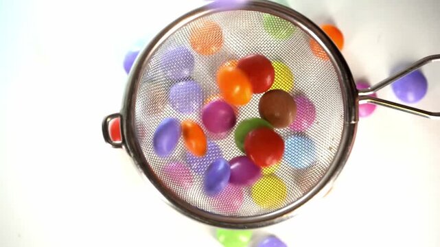 Close-up of a hand using a metal mesh strainer to scoop and manipulate colorful candy-coated chocolate drops on a clean white background, perfect for culinary and food-related concepts.