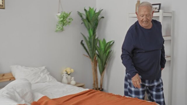 Man making bed and arranging pillow with orange blanket by bedside shelf and plants in a bedroom inside a building; morning routine calm.