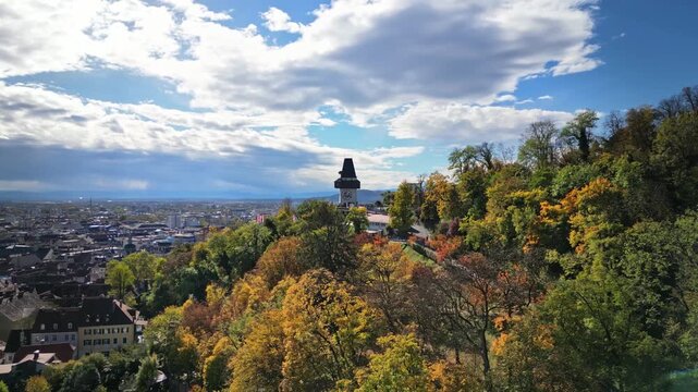 Aerial view of Schlossberg hill with the clock tower surrounded by autumn trees overlooking the city skyline in Graz, Steiermark, Austria.