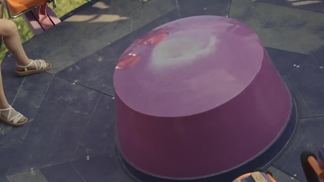 Close-up view of a glossy purple part of an amusement park ride. The smooth, reflective surface catches sunlight, creating a playful atmosphere. Nearby, glimpses of sandals and ride seating hint at ch