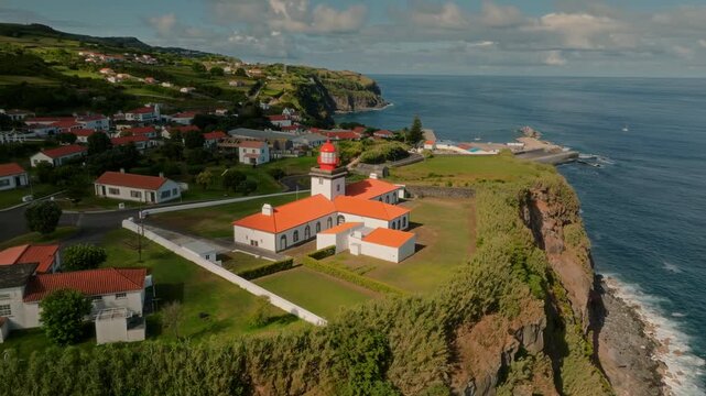 Aerial drone pan shot of the Farol de Lajes lighthouse with its distinct red roof, situated on the rugged coastline of Lajes das Flores, Azores.