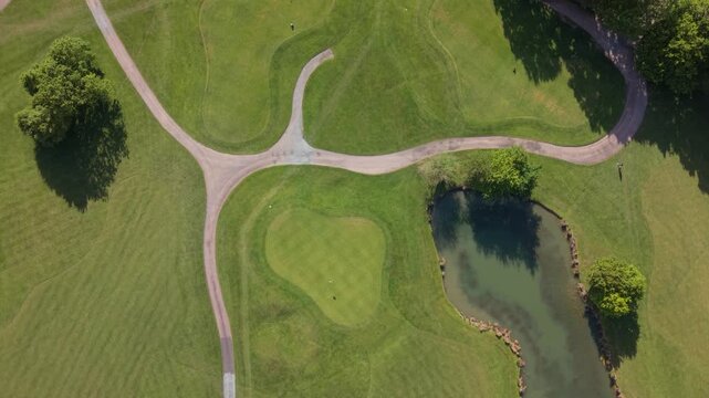Sunny daytime aerial view of a well-kept golf course. Lush green fairways, putting greens, and a water hazard are connected by winding cart paths. Trees cast long shadows.