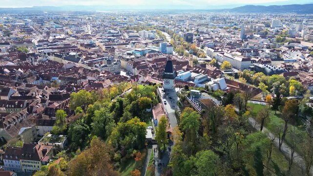 Aerial view of the Uhrturm clock tower on Schlossberg hill surrounded by autumn trees and the historic city skyline in Graz, Styria, Austria.