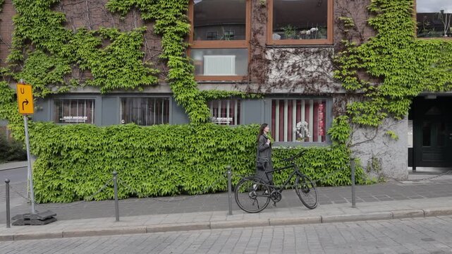 Woman walks past ivy-covered building with parked bicycle on cobblestone street