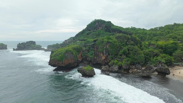 Aerial view of Siung Beach featuring lush green cliffs, white sand, and crashing ocean waves in Tepus, Special Region of Yogyakarta, Indonesia.